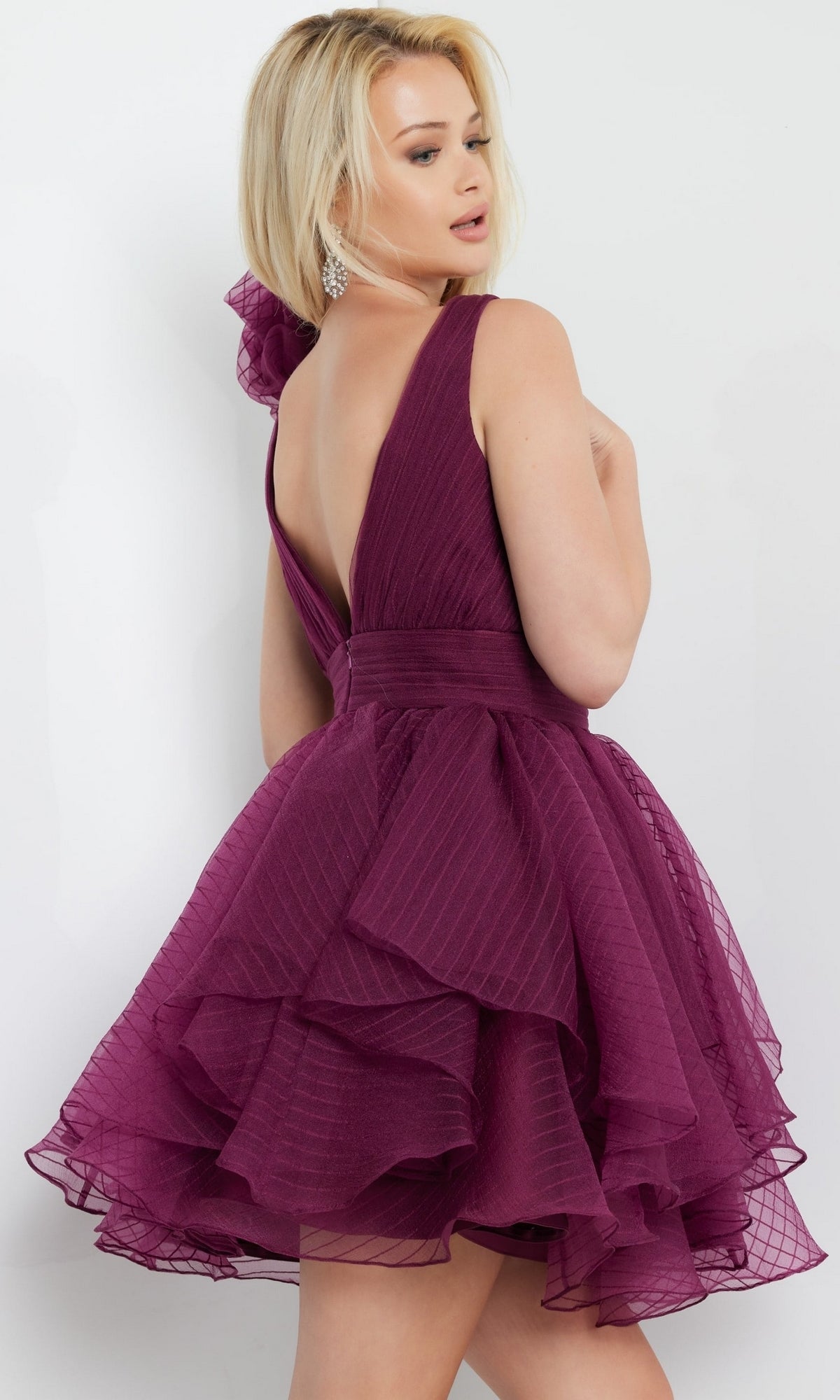 Woman wears the Jovani Rosette-Shoulder Short Party Dress 22779, featuring a sleeveless design, deep v-back, ruffled skirt, and textured magenta fabric as she poses facing backward against a plain light background.